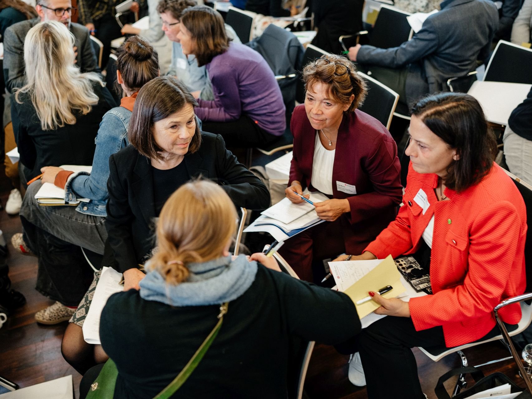 Vier Frauen sitzen in einem Stuhlkreis zusammen und unterhalten sich, im Hintergrund sind mehr Menschen im Gespräch in Kleingruppen zu sehen.