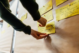 Eine Mitarbeiterin der Geschäftsstelle pinnt eine Karteikarte an eine Pinnwand, an der bereits weitere Karten hängen.
