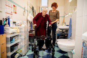 Eine Pflegerin unterstützt einer Seniorin mit Rollator im Bad