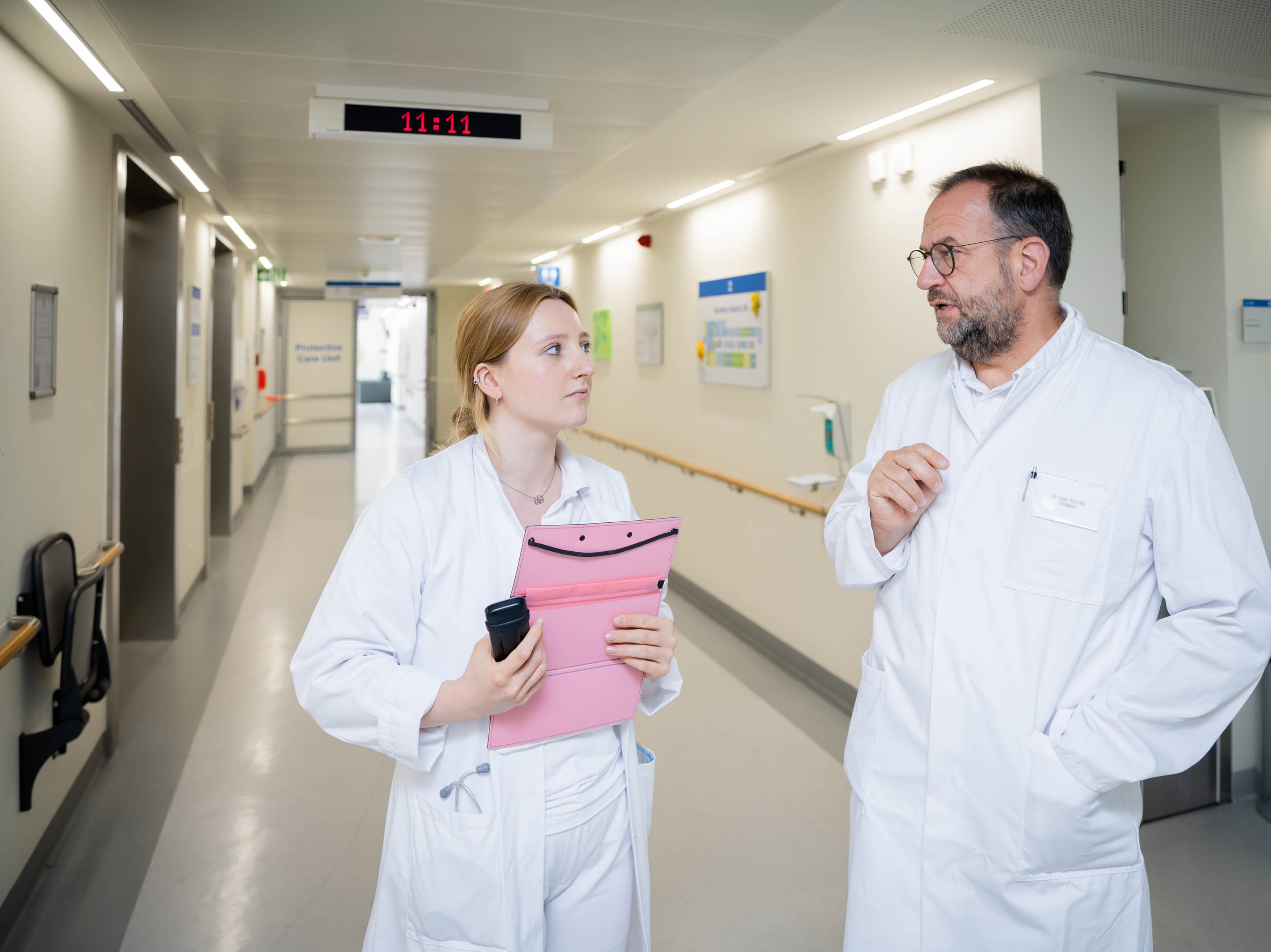 Zwei medizinische Fachkräfte in weißen Kitteln stehen im Krankenhausflur und sprechen miteinander; eine Person hält eine pinke Mappe.