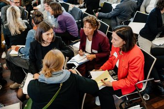 Vier Frauen sitzen in einem Stuhlkreis zusammen und unterhalten sich, im Hintergrund sind mehr Menschen im Gespräch in Kleingruppen zu sehen.