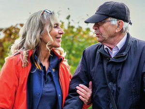 Eine Frau und ein älterer Mann gehen im Freien nebeneinander. Die Frau lächelt den Mann an und berührt unterstützend seinen Arm, während er einen Rollator schiebt. Unter dem Bild steht der Text „Modellprojekt SABD-Fam – Teilprojekt 1: Entwicklung und Umsetzung von SABD-Fam“.