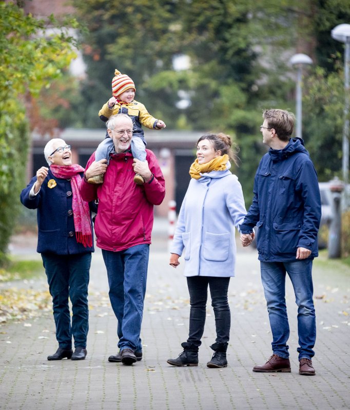 Junges Elternpaar und Großelternpaar beim gemeinsamen Spaziergang. Auf den Schultern des Großvaters sitzt ein lachendes Kleinkind.