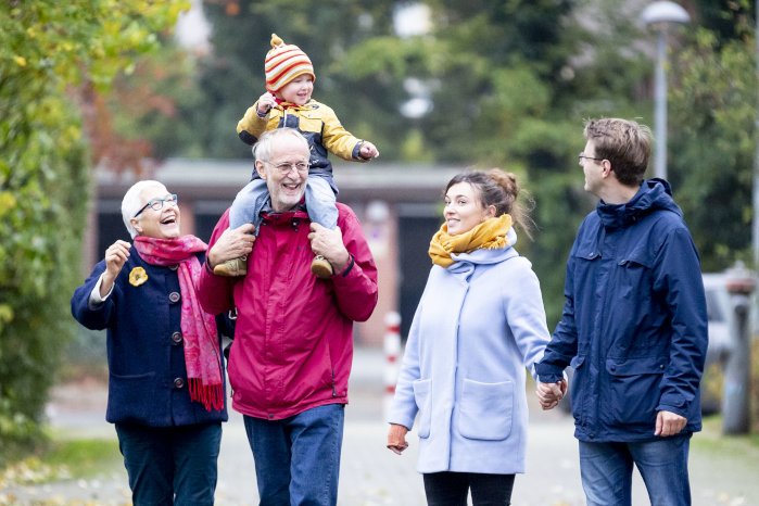 Junges Elternpaar und Großelternpaar beim gemeinsamen Spaziergang. Auf den Schultern des Großvaters sitzt ein lachendes Kleinkind.