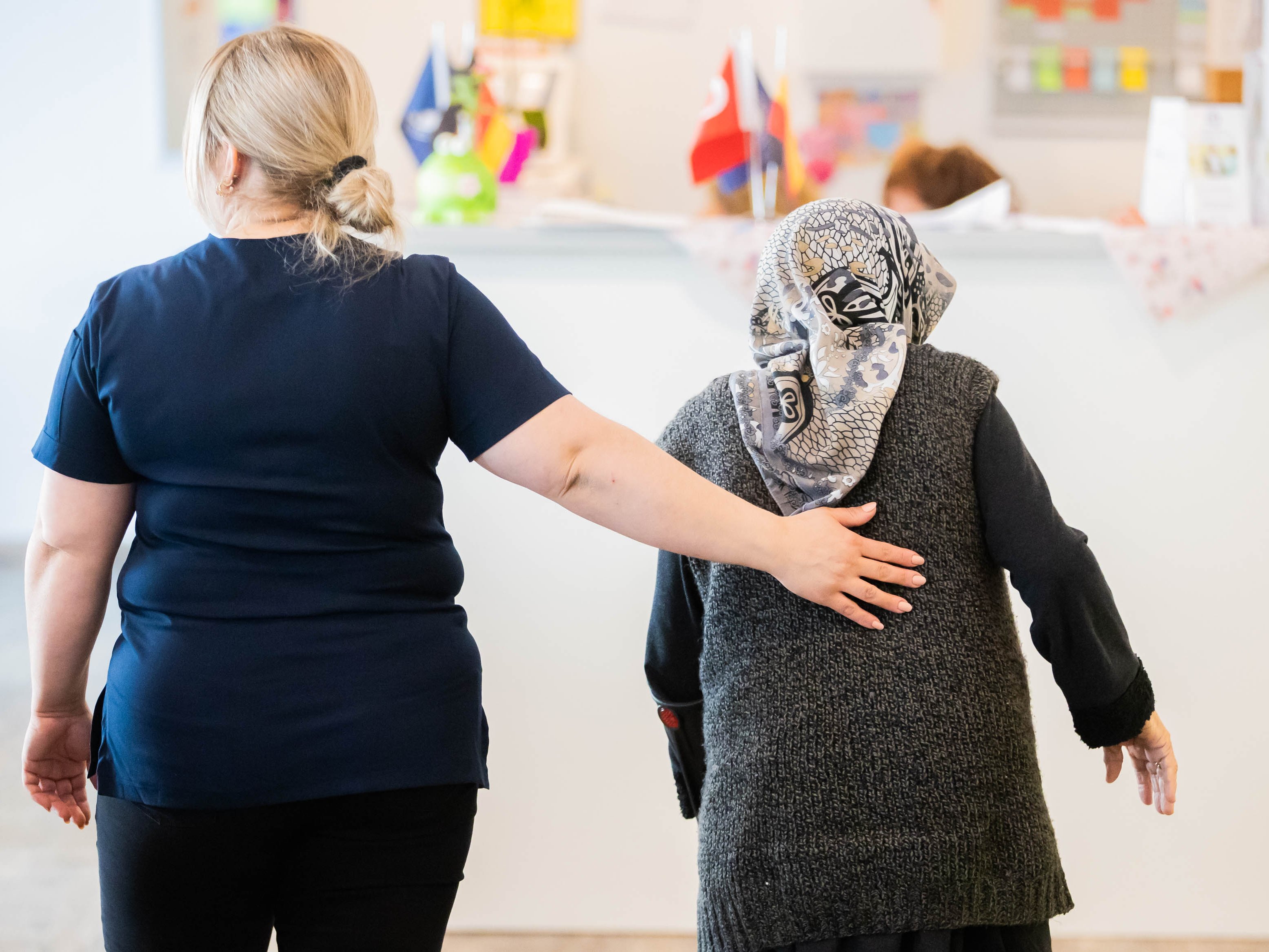 Eine Pflegefachperson läuft neben einer älteren Person, beide sind von hinten zu sehen. ©Christoph Soeder/DZA