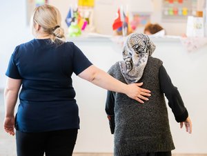 Eine Pflegefachperson läuft neben einer älteren Person, beide sind von hinten zu sehen. ©Christoph Soeder/DZA