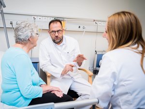 Ein Arzt spricht in einem Krankenhauszimmer ruhig und erklärend mit einer älteren Patientin, während eine weitere medizinische Fachkraft aufmerksam zuhört.