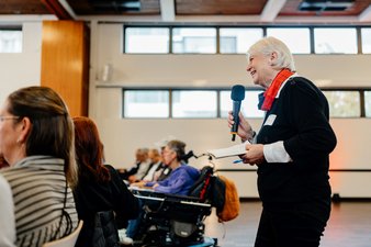 Brigitte Bührlen (Wir! Stiftung pflegender Angehöriger) steht hinter den Stuhlreihen des Plenums und stellt eine Rückfrage zum Vortrag von Dr. Christine Schwendner..