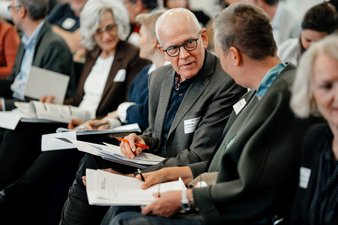 Die Teilnhemenden sitzen im Plenum, haben ein Konzeptpapier auf dem Schoß und sprechen paarweise miteinander. 