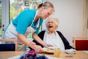 Eine Pflegekraft stellt einen Teller vor einer älteren Frau auf den Tisch