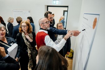 Personen stehen im Tagungsraum vor einem Poster. Zwei Teilnehmer sind im Gespräch zum Posterinhalt, einer zeigt mit dem Stift auf eine bestimmte Stelle des Posters, der zweite mit dem Zeigefinger.