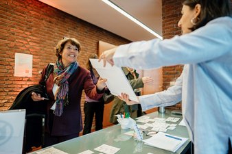 Willkommen auf der Netzwerktagung: Eine Teilnehmerin erhält einen Umschlag mit Tagungsmaterial von einer Mitarbeierin der Geschäftsstelle Nationale Demenzstrategie.