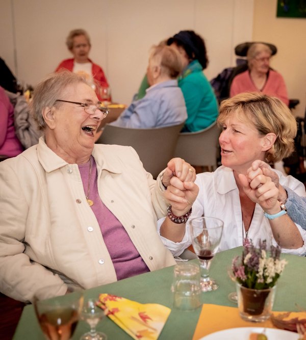 Festliche Veranstaltung, Tisch mit zwei gut gelaunten Seniorinnen, dazwischen eine jüngere Frau in der Hocke zwischen den beiden älteren Frauen, die die beiden lachend anspricht.