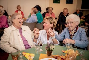 Festliche Veranstaltung, Tisch mit zwei gut gelaunten Seniorinnen, dazwischen eine jüngere Frau in der Hocke zwischen den beiden älteren Frauen, die die beiden lachend anspricht.