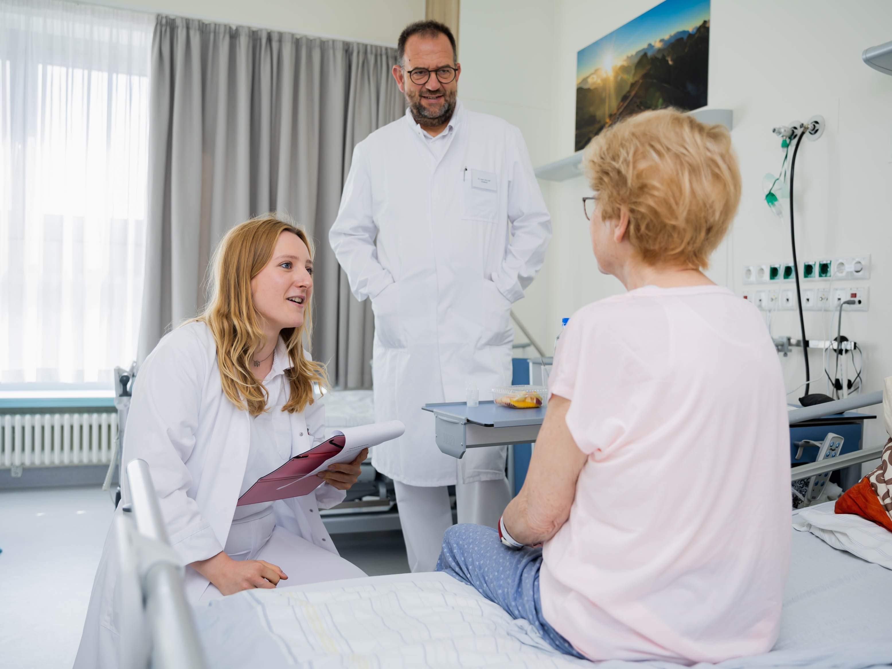 Eine Ärztin und ein Arzt sprechen mit einer Frau, die auf einem Krankenhausbett sitzt.