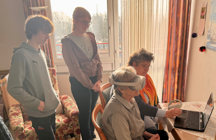 Jugendliche zeigen einer älteren Frau mit Demenz ihr Projekt "Virtual Reality gegen Demenz", die ältere Dame mit einer Begleitperson an einem Laptop und hat eine VR-Brille auf.