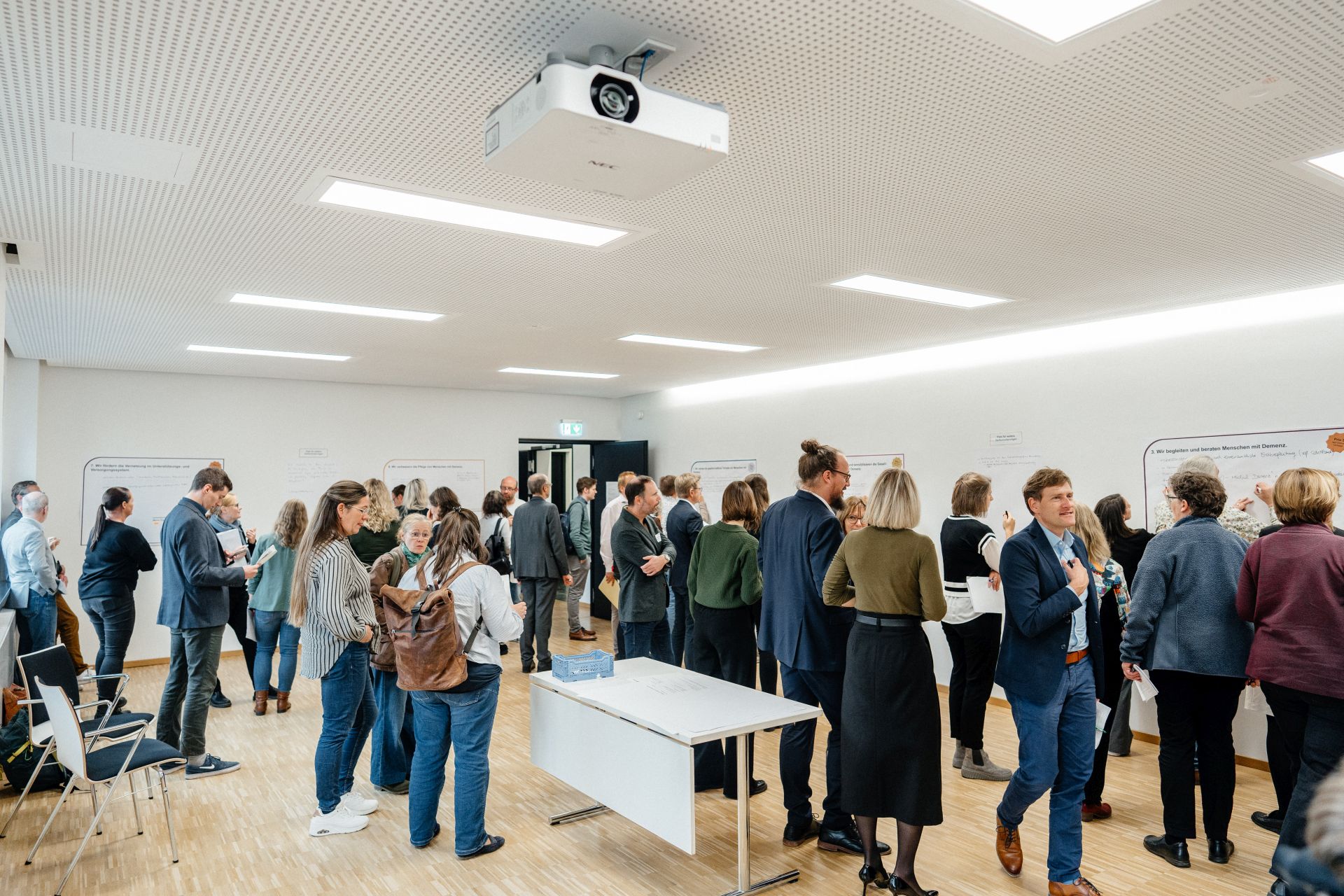 Mehrere Personen stehen in kleinen Gruppen in einem hellen Tagungsraum und diskutieren vor großen Wandpostern. Einige schreiben Notizen, andere sprechen miteinander oder lesen die Beiträge. Die Szene zeigt eine aktive Arbeitsphase im Rahmen der Netzwerktagung Nationale Demenzstrategie.